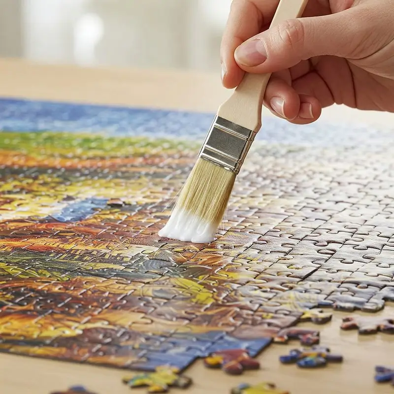 Eine Nahaufnahme einer Hand, die mit einem breiten Pinsel weißen Kleber auf ein fertig zusammengesetztes Puzzle aufträgt. Das Puzzle zeigt ein farbenfrohes Motiv, und der Kleber verleiht ihm einen glänzenden Effekt.